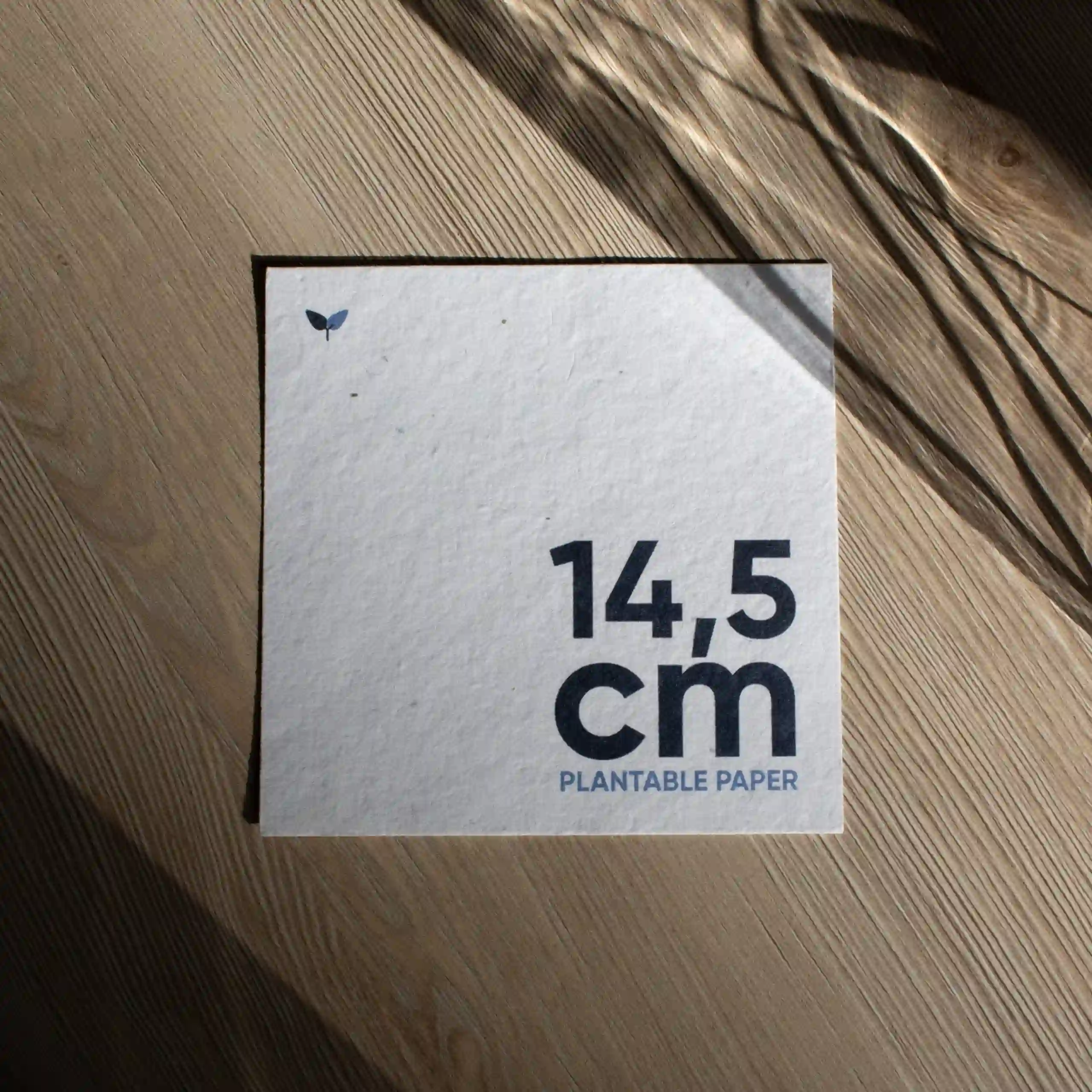 Tarjeta cuadrada impresa en papel plantable sobre una superficie de madera con sombras