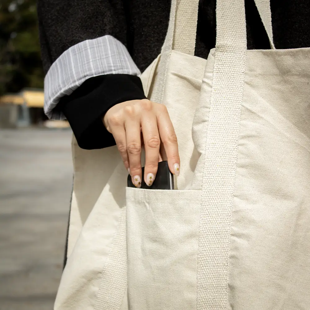 Una mano guardando un móvil en el bolsillo exterior de una tote bag de algodón