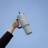 Mano alzando un vaso térmico gris de acero inoxidable con pajita con el cielo de fondo