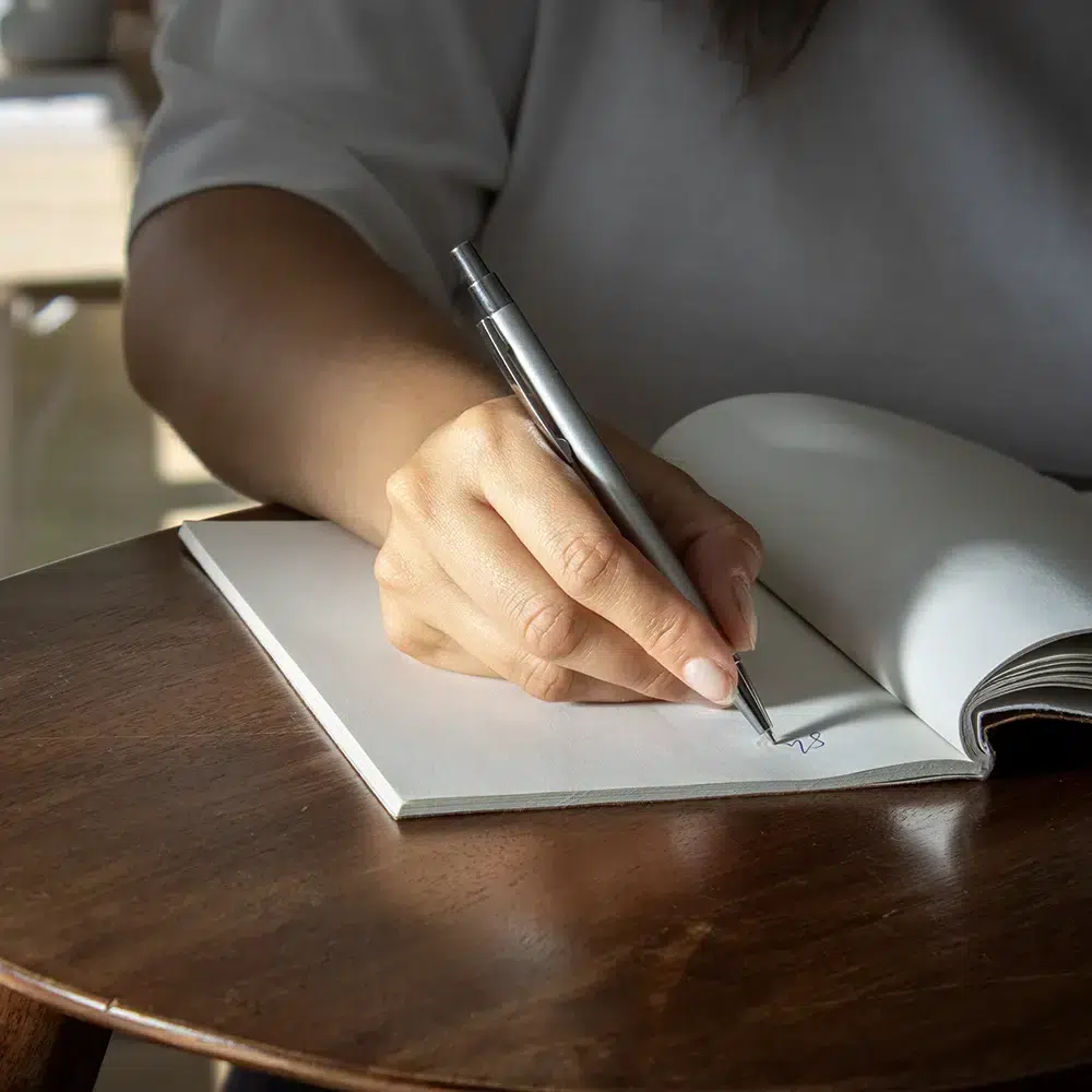 Mano escribiendo sobre un cuaderno con bolígrafo plateado de aluminio reciclado sobre una superficie de madera