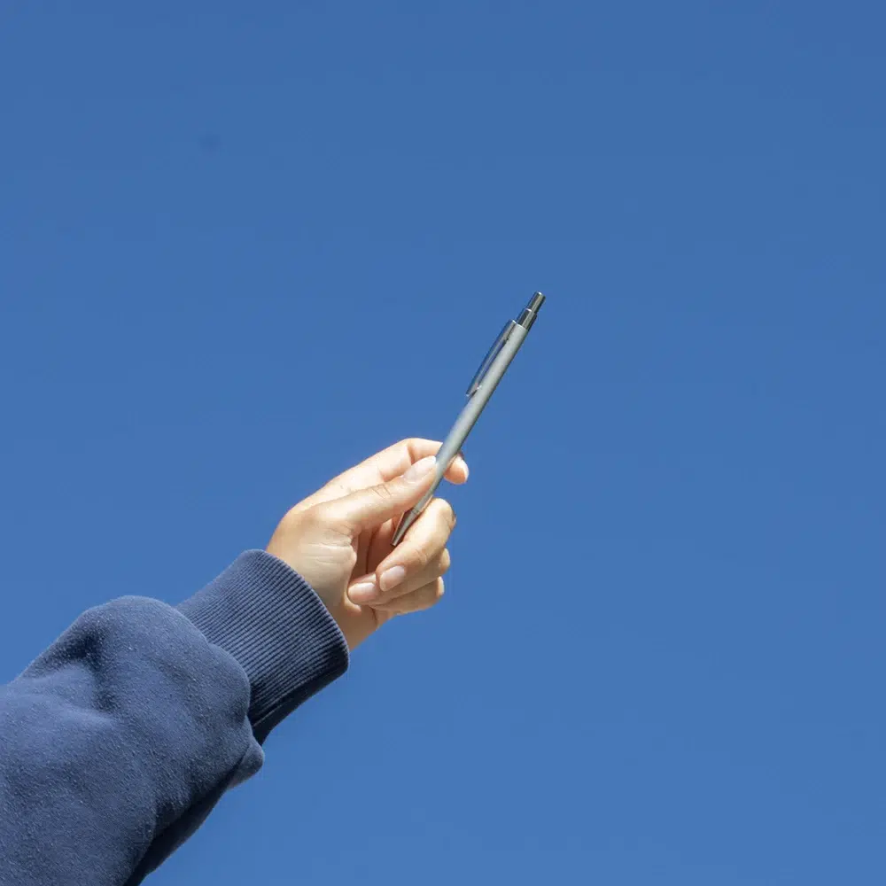 Bolígrafo de aluminio reciclado GANTE Mano alzando un bolígrafo plateado de aluminio reciclado con el cielo de fondo
