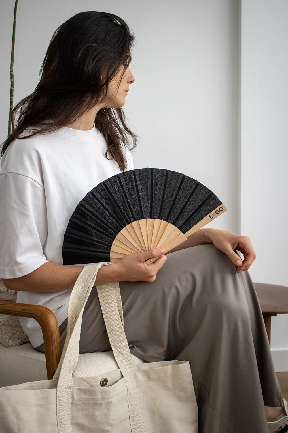Chica de perfil sujetando abanico negro con varillas de madera y tela poliéster abierto junto a una tote bag
