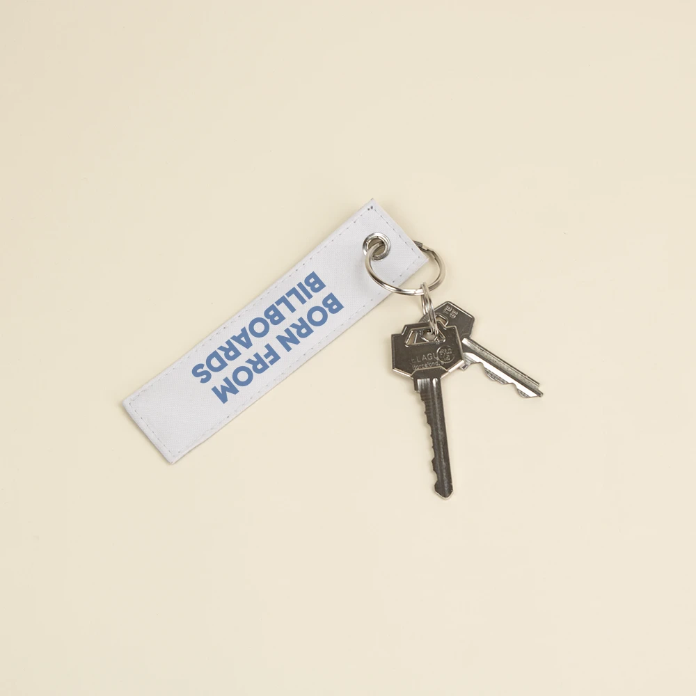 Llavero hecho con lona reciclada blanca personalizado con el texto impreso en azul: "Born from billboards" con dos llaves enganchadas de su anilla metálica