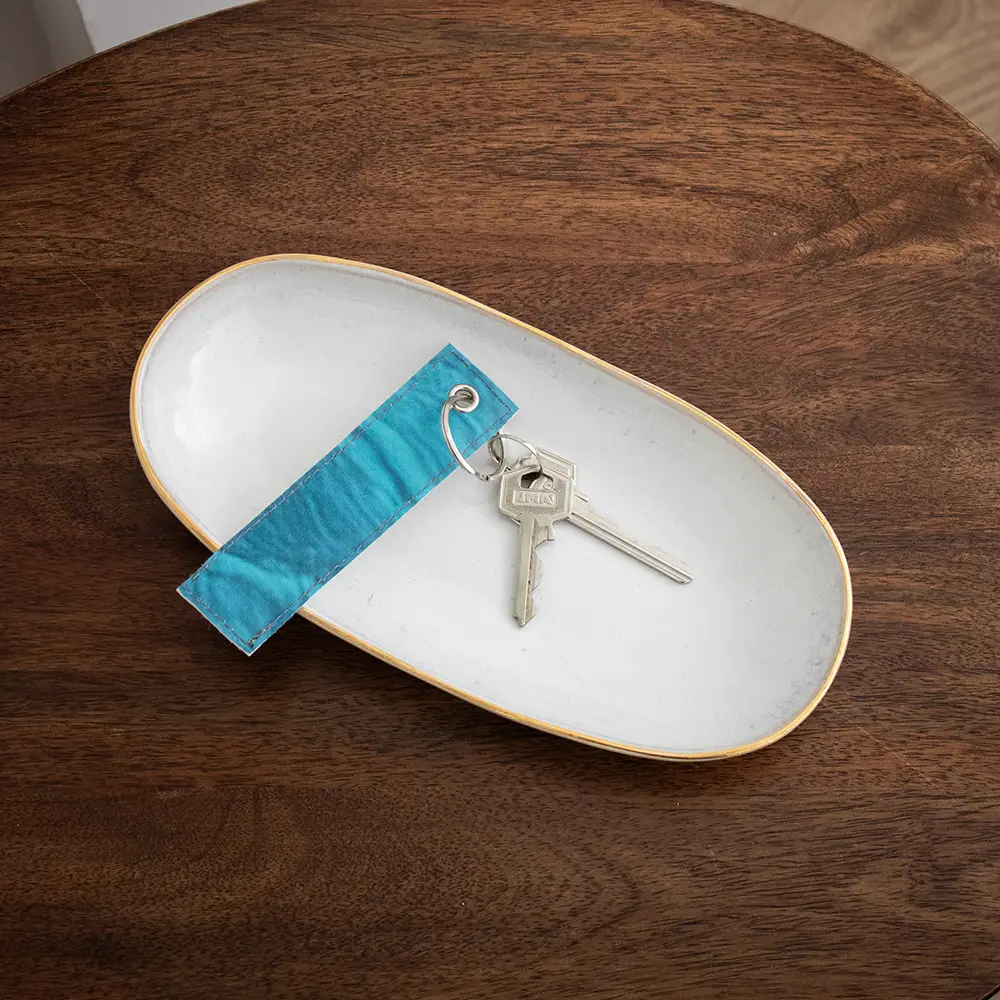 Llavero hecho con lona reciclada azul con dos llaves enganchadas de su anilla metálica, colocado sobre una bandeja blanca en una superficie de madera