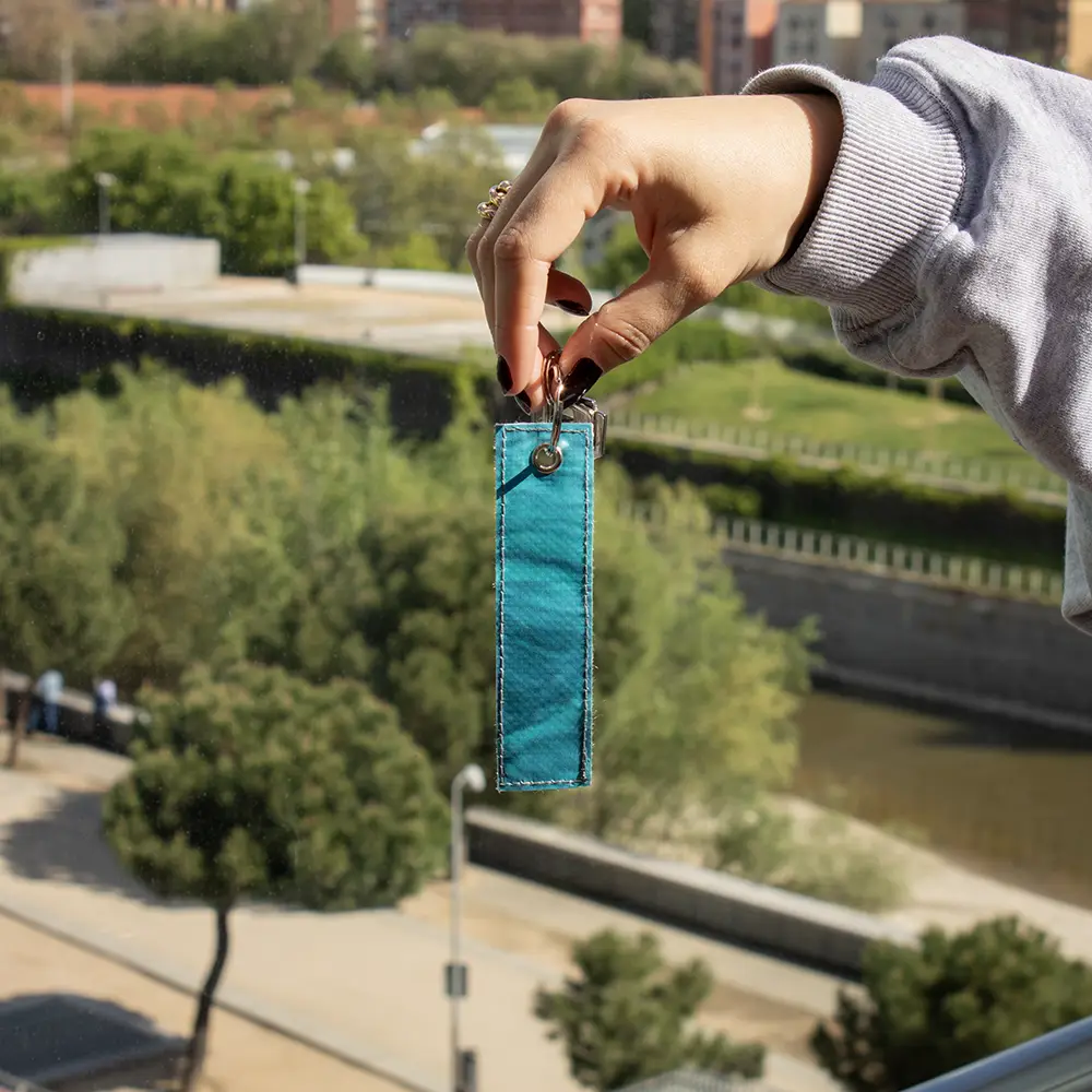 Mano mostrando un llavero azul hecho con lona reciclada, con la ciudad de fondo