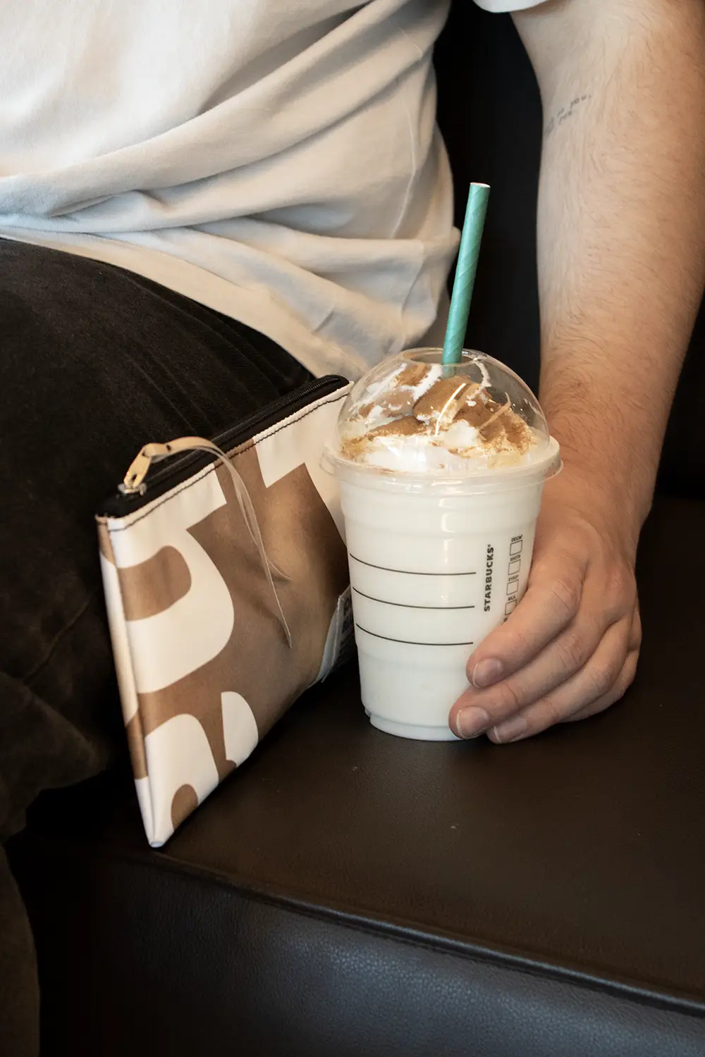 Mano sujetando un frappuccino junto a un estuche de lona reciclada de color marrón y blanco