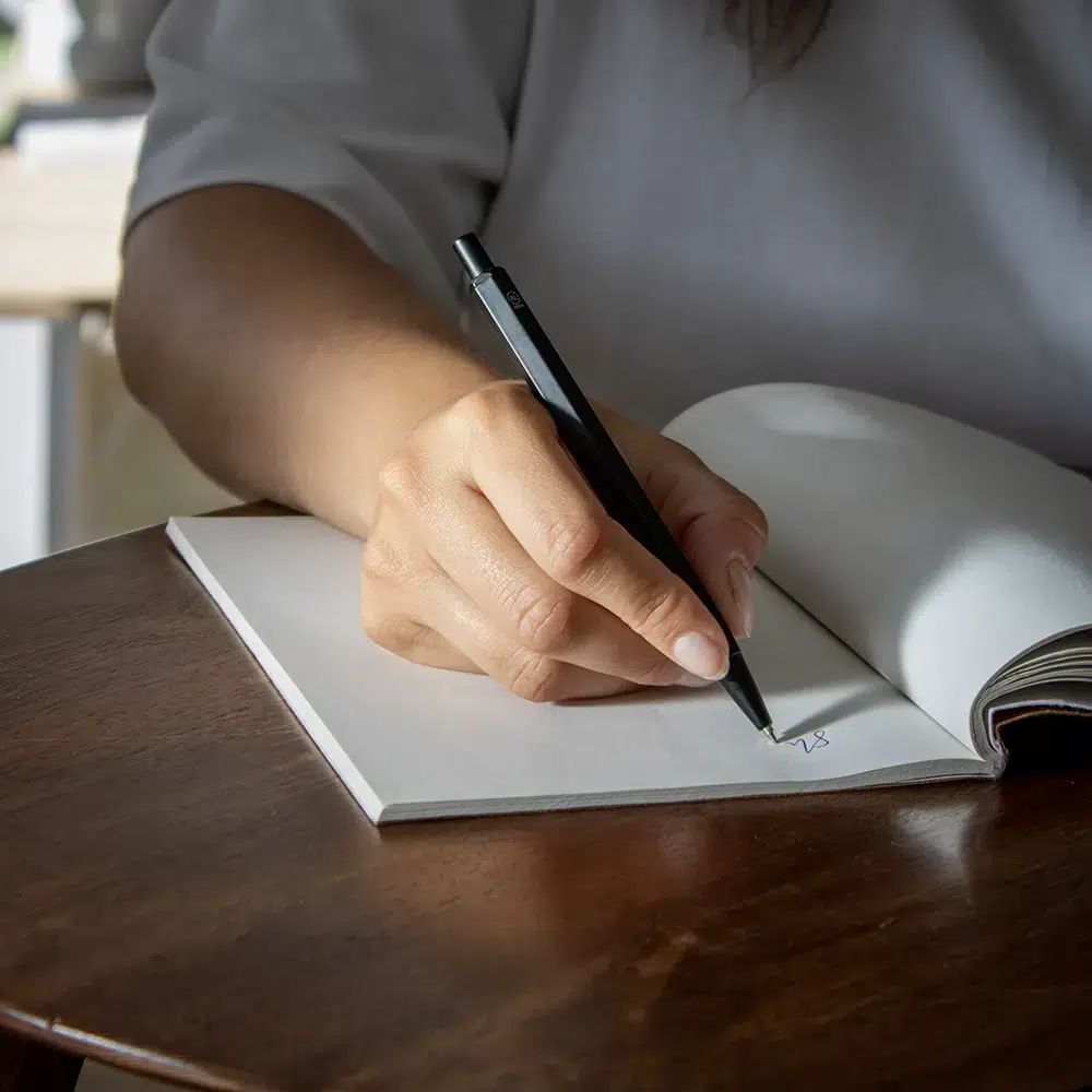 Mano escribiendo sobre un cuaderno con bolígrafo de aluminio negro sobre una superficie de madera