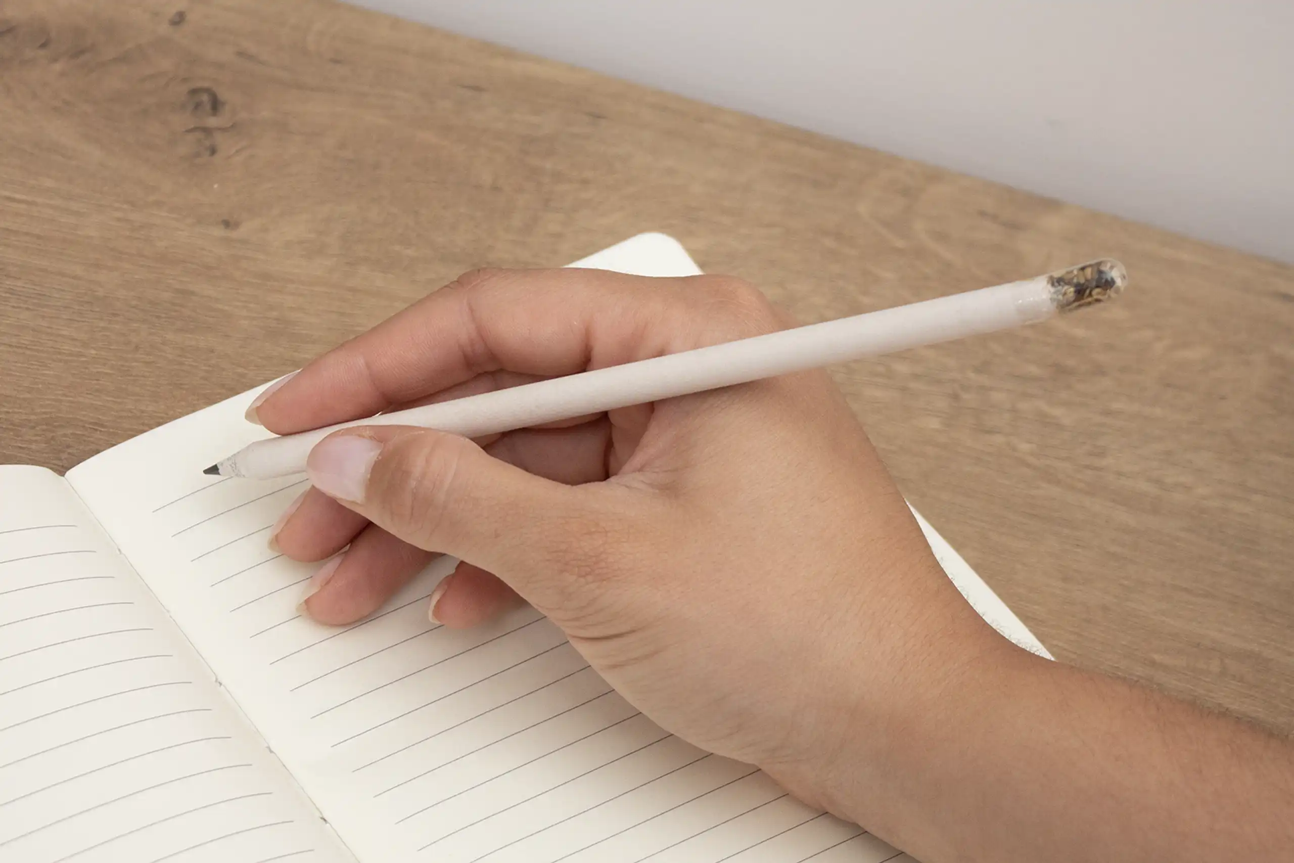 Mano escribiendo con un lápiz plantable blanco en un cuaderno abierto sobre una superficie de madera
