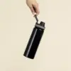 Mano sosteniendo una botella deportiva negra personalizada en aluminio reciclado sobre un fondo beige