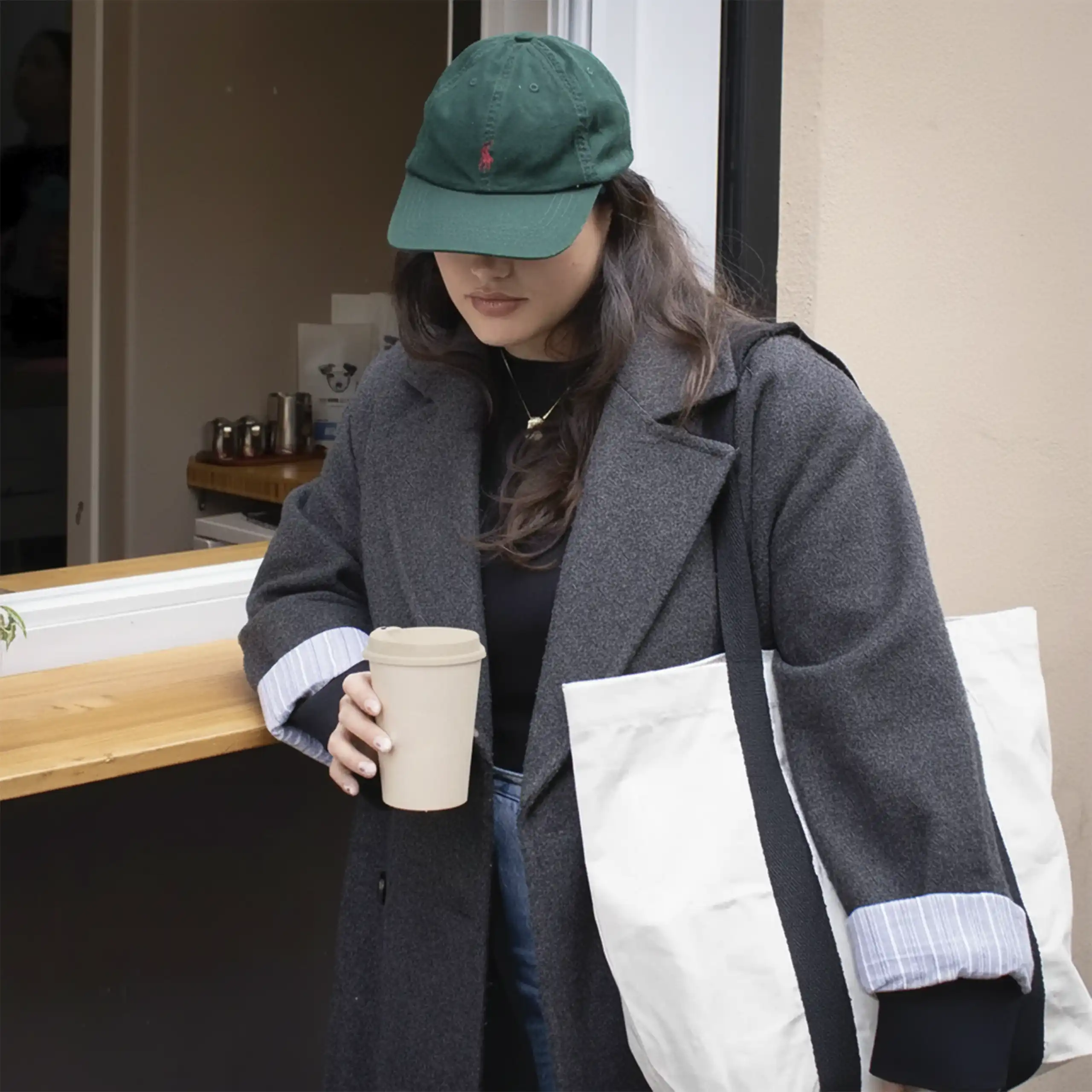 Chica con una gorra verde apoyada en la barra de una cafetería llevando un vaso take away y una tote bag de algodón blanca
